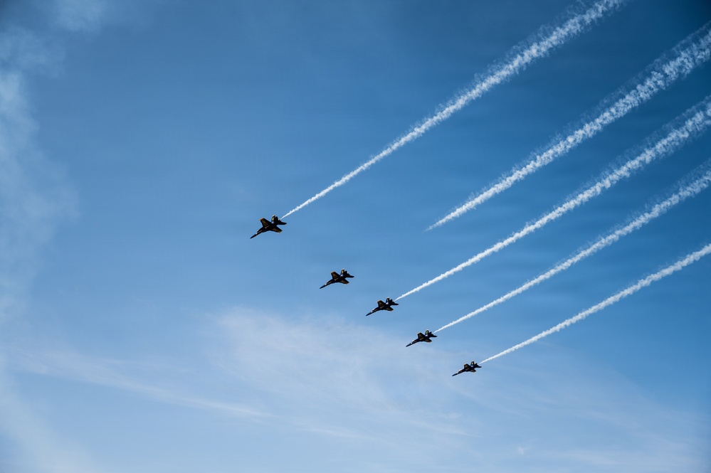 Six military jet aircraft are flying in a diagonal echelon formation against a clear blue sky, each producing white contrails that extend toward the upper right of the frame. The aircraft appear to be performing a precision formation flight, consistent with an airshow or ceremonial flyover. The silhouetted jets and parallel contrail lines create a strong visual pattern across the image.