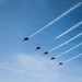 Six military jet aircraft are flying in a diagonal echelon formation against a clear blue sky, each producing white contrails that extend toward the upper right of the frame. The aircraft appear to be performing a precision formation flight, consistent with an airshow or ceremonial flyover. The silhouetted jets and parallel contrail lines create a strong visual pattern across the image.