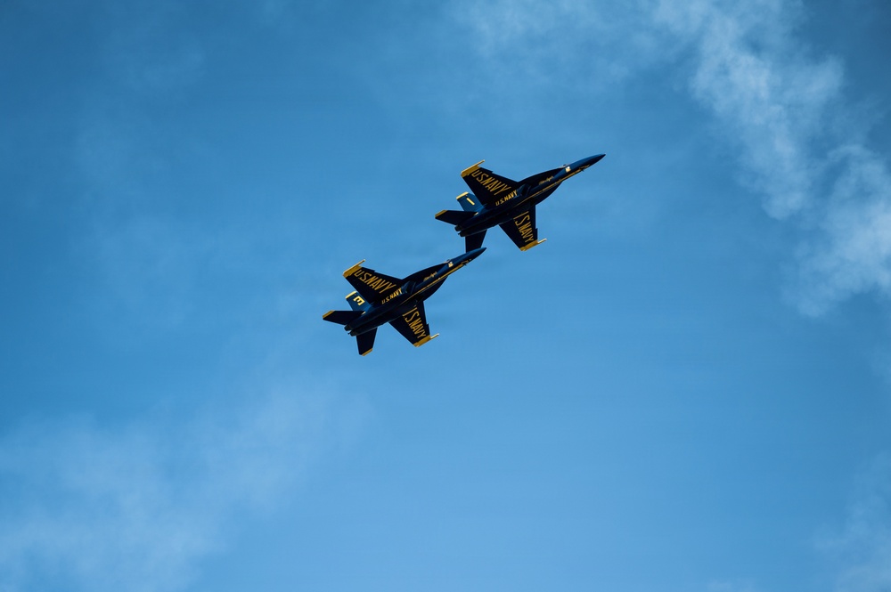 Two U.S. Navy Blue Angels F/A-18 Hornet jets are captured in close formation flight against a predominantly blue sky with light cloud cover. The aircraft are painted in the iconic dark blue and gold color scheme with 'U.S. NAVY' and 'U.S. NAVY Blue Angels' markings clearly visible. The jets appear to be performing a precision formation maneuver, flying in tight parallel alignment.