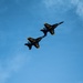 Two U.S. Navy Blue Angels F/A-18 Hornet jets are captured in close formation flight against a predominantly blue sky with light cloud cover. The aircraft are painted in the iconic dark blue and gold color scheme with 'U.S. NAVY' and 'U.S. NAVY Blue Angels' markings clearly visible. The jets appear to be performing a precision formation maneuver, flying in tight parallel alignment.