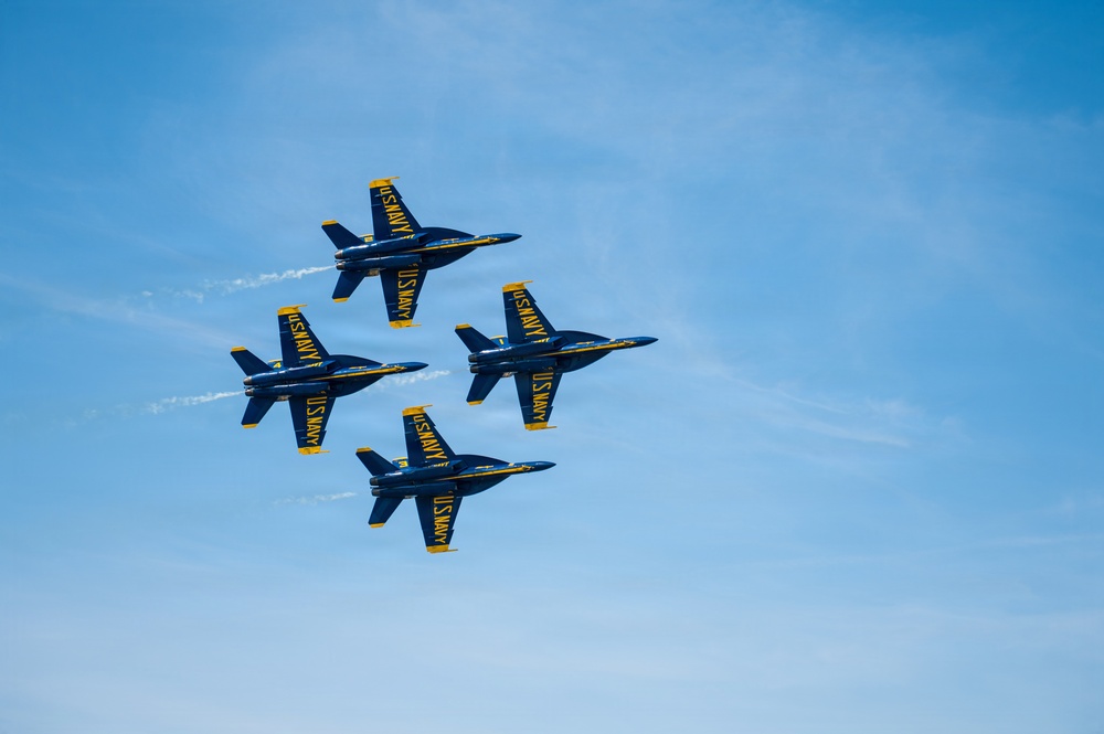 Four U.S. Navy Blue Angels aircraft are flying in a tight diamond formation against a clear blue sky. The jets are painted in their iconic blue and gold color scheme with 'U.S. NAVY' markings clearly visible on the fuselages and tail fins. This appears to be an airshow performance demonstrating precision formation flying.