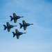 Four U.S. Navy Blue Angels aircraft are flying in a tight diamond formation against a clear blue sky. The jets are painted in their iconic blue and gold color scheme with 'U.S. NAVY' markings clearly visible on the fuselages and tail fins. This appears to be an airshow performance demonstrating precision formation flying.