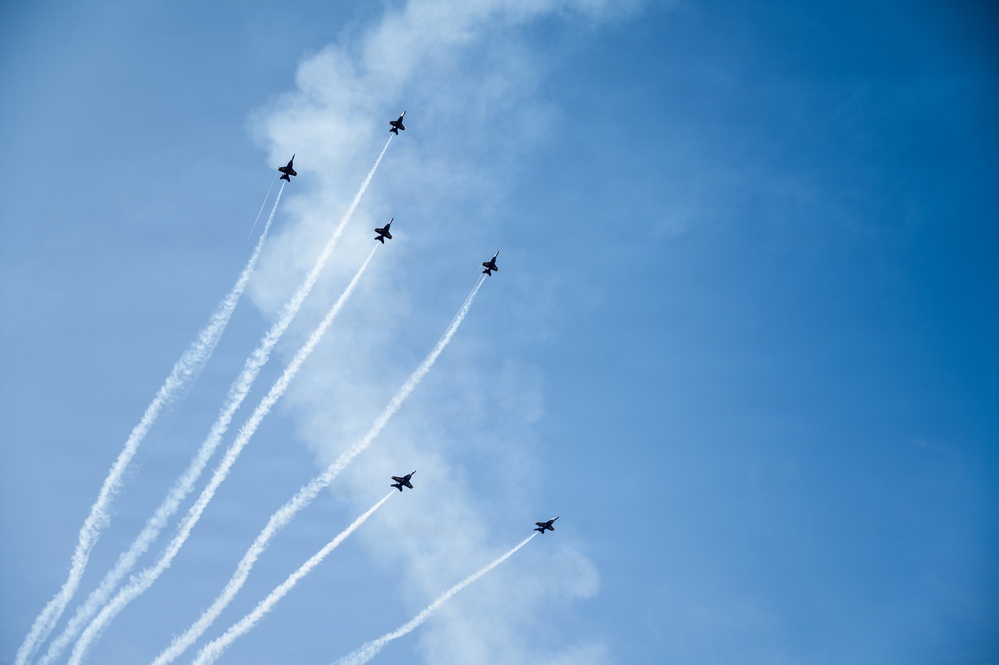 Six military jet aircraft are performing an aerial formation display against a blue sky with light cloud cover. The jets are flying in a diagonal echelon formation, each producing white contrails/smoke trails that extend downward to the lower left of the frame. The image captures a precision aerobatics demonstration, consistent with teams such as the U.S. Air Force Thunderbirds or U.S. Navy Blue Angels.