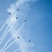 Six military jet aircraft are performing an aerial formation display against a blue sky with light cloud cover. The jets are flying in a diagonal echelon formation, each producing white contrails/smoke trails that extend downward to the lower left of the frame. The image captures a precision aerobatics demonstration, consistent with teams such as the U.S. Air Force Thunderbirds or U.S. Navy Blue Angels.