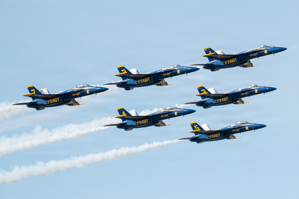 Six U.S. Navy Blue Angels aircraft are flying in a tight diamond formation against a clear blue sky. The jets are painted in their iconic blue and gold color scheme with 'U.S. NAVY' and 'Blue Angels' markings clearly visible. Several of the aircraft are producing white smoke trails, indicating an active airshow performance.