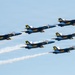 Six U.S. Navy Blue Angels aircraft are flying in a tight diamond formation against a clear blue sky. The jets are painted in their iconic blue and gold color scheme with 'U.S. NAVY' and 'Blue Angels' markings clearly visible. Several of the aircraft are producing white smoke trails, indicating an active airshow performance.