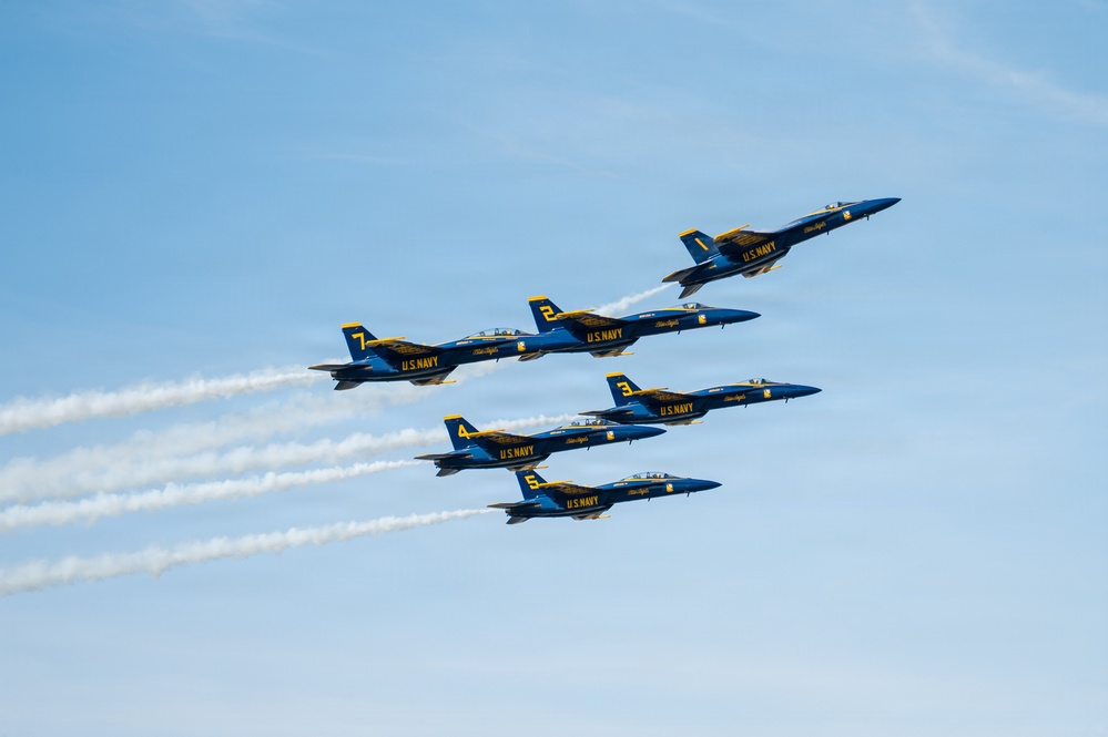 Six U.S. Navy Blue Angels aircraft are flying in a tight diamond/delta formation against a clear blue sky. The jets, numbered 1 through 5 and 7, are painted in their signature blue and gold livery with 'U.S. NAVY' and 'Blue Angels' markings. Several of the aircraft are emitting white smoke trails as they perform their precision aerobatic display.