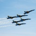 Six U.S. Navy Blue Angels aircraft are flying in a tight diamond/delta formation against a clear blue sky. The jets, numbered 1 through 5 and 7, are painted in their signature blue and gold livery with 'U.S. NAVY' and 'Blue Angels' markings. Several of the aircraft are emitting white smoke trails as they perform their precision aerobatic display.