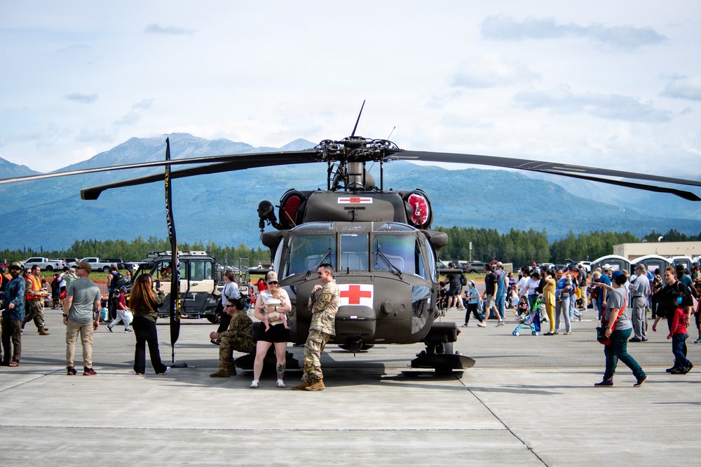 A military medical helicopter, identified by the Red Cross markings, is on static display at what appears to be a public airshow or open house event at a military airfield. Crowds of civilians, including families with children, are gathered around the aircraft on the tarmac. Mountain ranges and forested terrain visible in the background suggest a Pacific Northwest or Alaskan location.
