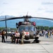 A military medical helicopter, identified by the Red Cross markings, is on static display at what appears to be a public airshow or open house event at a military airfield. Crowds of civilians, including families with children, are gathered around the aircraft on the tarmac. Mountain ranges and forested terrain visible in the background suggest a Pacific Northwest or Alaskan location.