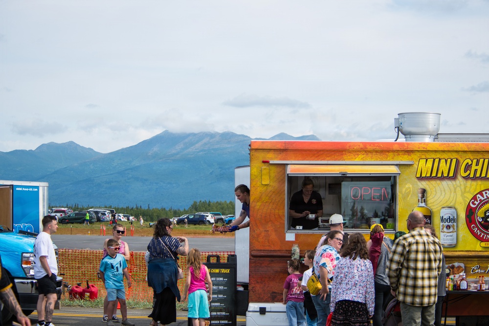 A busy food truck scene at what appears to be an outdoor event or fair, with a yellow-orange 'Mini Chi' (likely Mini Chimichanga) truck serving customers. A crowd of adults and children are gathered around the truck, which displays an 'OPEN' neon sign and Modelo beer branding. The backdrop features a dramatic mountain range under a partly cloudy sky, suggesting a location in Alaska or the Pacific Northwest.