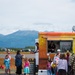 A busy food truck scene at what appears to be an outdoor event or fair, with a yellow-orange 'Mini Chi' (likely Mini Chimichanga) truck serving customers. A crowd of adults and children are gathered around the truck, which displays an 'OPEN' neon sign and Modelo beer branding. The backdrop features a dramatic mountain range under a partly cloudy sky, suggesting a location in Alaska or the Pacific Northwest.