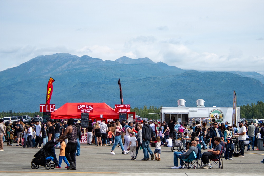 An outdoor food festival or fair taking place on a large paved surface, likely an airfield or parking lot, with a large crowd of attendees browsing food vendors. Red canopy tents and food trucks are visible offering items such as 'Cowboy Fries,' 'T-Legs,' and 'Pulled Pork Sandwich.' A dramatic mountain range and forested treeline form the backdrop under a partly cloudy sky, suggesting a location in Alaska or the Pacific Northwest.