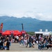 An outdoor food festival or fair taking place on a large paved surface, likely an airfield or parking lot, with a large crowd of attendees browsing food vendors. Red canopy tents and food trucks are visible offering items such as 'Cowboy Fries,' 'T-Legs,' and 'Pulled Pork Sandwich.' A dramatic mountain range and forested treeline form the backdrop under a partly cloudy sky, suggesting a location in Alaska or the Pacific Northwest.