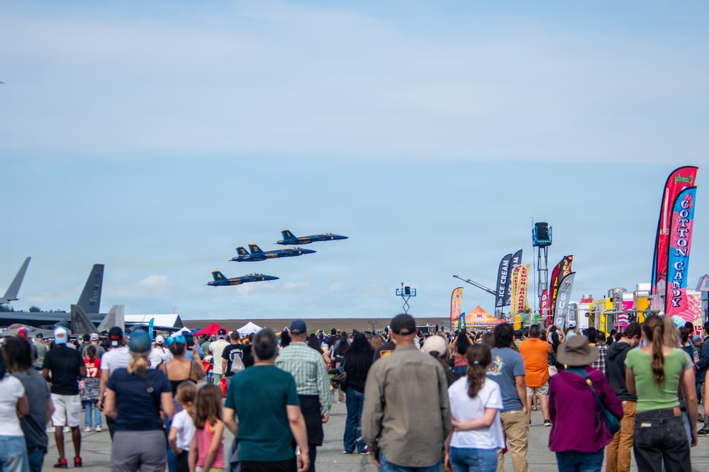 An air show is taking place at a military or civilian airfield, with a large crowd of spectators watching three blue military jet aircraft flying in formation overhead. The event features vendor booths and food stalls along the right side, including signs for ice cream and cotton candy. The scene captures a festive public event with hundreds of attendees gathered on the tarmac.