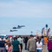 An air show is taking place at a military or civilian airfield, with a large crowd of spectators watching three blue military jet aircraft flying in formation overhead. The event features vendor booths and food stalls along the right side, including signs for ice cream and cotton candy. The scene captures a festive public event with hundreds of attendees gathered on the tarmac.