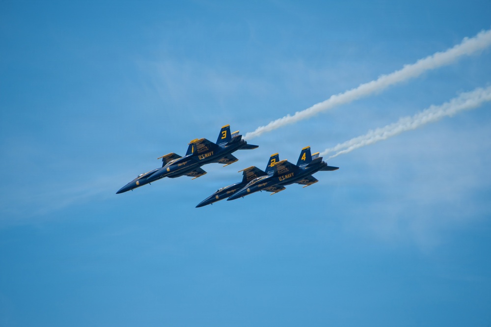 Four U.S. Navy Blue Angels F/A-18 Hornet jets fly in tight diamond formation against a clear blue sky. The aircraft are numbered 1, 2, 3, and 4, painted in their signature blue and gold livery. At least one jet is producing a white smoke trail extending toward the upper right of the frame.