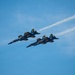 Four U.S. Navy Blue Angels F/A-18 Hornet jets fly in tight diamond formation against a clear blue sky. The aircraft are numbered 1, 2, 3, and 4, painted in their signature blue and gold livery. At least one jet is producing a white smoke trail extending toward the upper right of the frame.