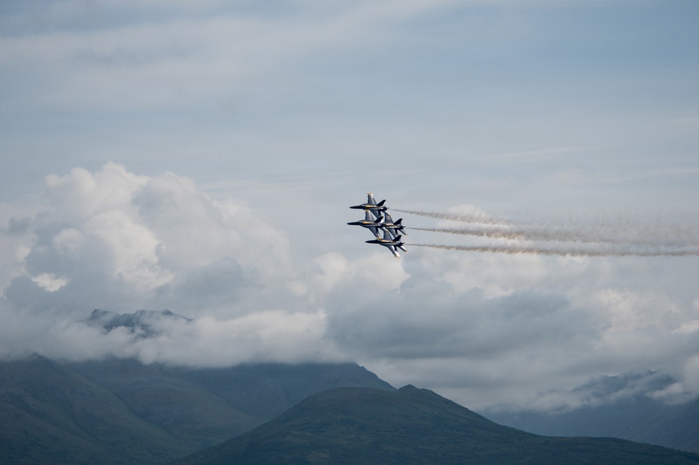 Four jet aircraft fly in tight diamond formation against a partly cloudy sky, leaving white smoke trails behind them. The aircraft appear to be a precision aerobatic demonstration team, likely the U.S. Navy Blue Angels based on their formation and coloring. Mountain peaks are visible in the lower portion of the frame beneath the cloud layer.