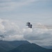 Four jet aircraft fly in tight diamond formation against a partly cloudy sky, leaving white smoke trails behind them. The aircraft appear to be a precision aerobatic demonstration team, likely the U.S. Navy Blue Angels based on their formation and coloring. Mountain peaks are visible in the lower portion of the frame beneath the cloud layer.