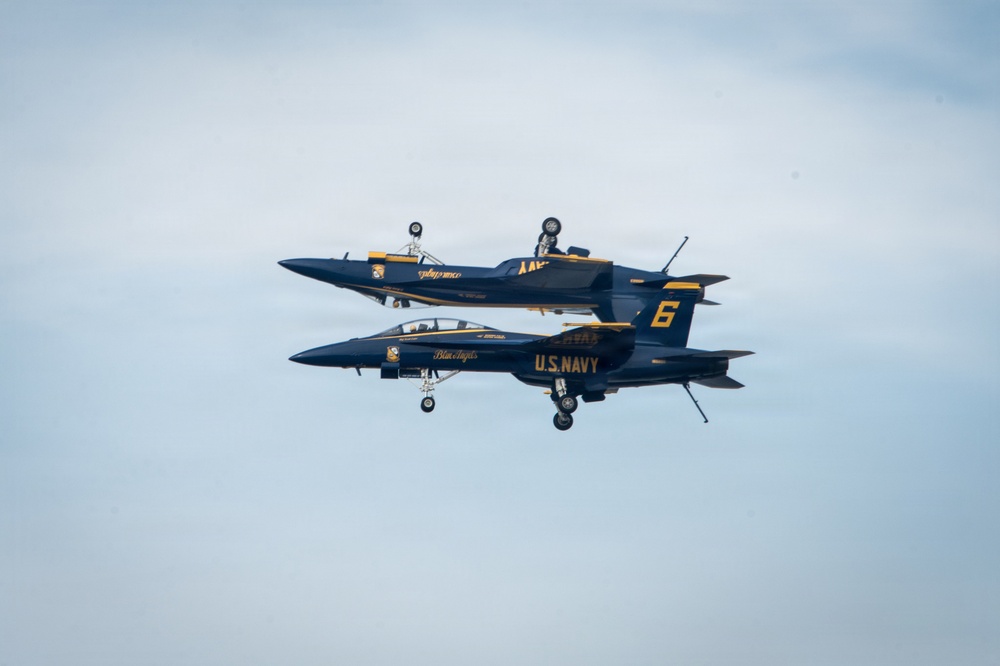 Two U.S. Navy Blue Angels F/A-18 Hornet jets are performing a mirror formation maneuver, with one aircraft flying inverted directly above the other. The jets are painted in the iconic navy blue and gold color scheme of the Blue Angels flight demonstration squadron. The aircraft are flying in extremely close proximity, showcasing precision aerobatic skill.