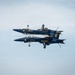 Two U.S. Navy Blue Angels F/A-18 Hornet jets are performing a mirror formation maneuver, with one aircraft flying inverted directly above the other. The jets are painted in the iconic navy blue and gold color scheme of the Blue Angels flight demonstration squadron. The aircraft are flying in extremely close proximity, showcasing precision aerobatic skill.