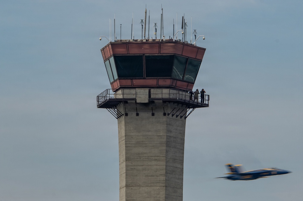 The image shows a concrete air traffic control tower with a reddish-brown glass cab at the top, featuring an exterior observation walkway where two people are standing. In the lower right corner, a blue and yellow military jet aircraft — consistent with the U.S. Navy Blue Angels livery — is captured in motion blur, indicating high speed flight. The overcast sky provides a neutral backdrop, emphasizing both the static tower and the dynamic aircraft.