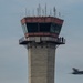 The image shows a concrete air traffic control tower with a reddish-brown glass cab at the top, featuring an exterior observation walkway where two people are standing. In the lower right corner, a blue and yellow military jet aircraft — consistent with the U.S. Navy Blue Angels livery — is captured in motion blur, indicating high speed flight. The overcast sky provides a neutral backdrop, emphasizing both the static tower and the dynamic aircraft.