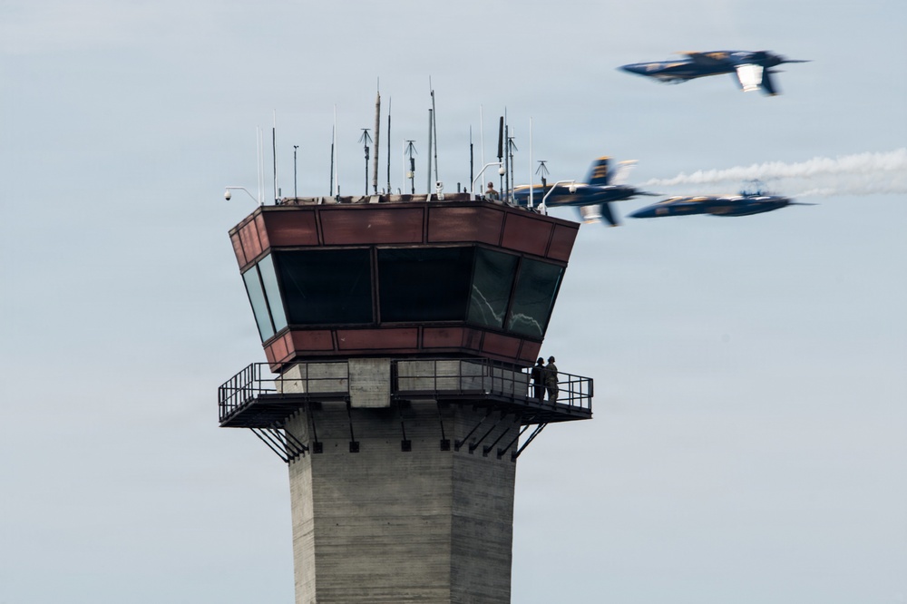 The image shows an air traffic control tower with two people standing on its observation balcony, watching a formation of military jet aircraft flying past. The jets, appearing to be Blue Angels based on their blue and yellow color scheme, are performing a close formation flyby with at least one aircraft producing a visible smoke trail. The scene is consistent with an airshow performance at a military or civilian airfield.