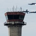 The image shows an air traffic control tower with two people standing on its observation balcony, watching a formation of military jet aircraft flying past. The jets, appearing to be Blue Angels based on their blue and yellow color scheme, are performing a close formation flyby with at least one aircraft producing a visible smoke trail. The scene is consistent with an airshow performance at a military or civilian airfield.
