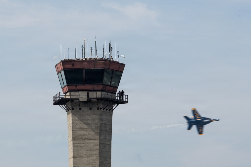 An air traffic control tower is prominently featured in the left portion of the image, with two silhouetted figures visible on its observation deck. A military jet aircraft, identifiable by its blue and yellow color scheme consistent with the U.S. Navy Blue Angels demonstration team, streaks past in the right portion of the frame with motion blur indicating high speed. The scene appears to be captured during an airshow event.