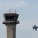 An air traffic control tower is prominently featured in the left portion of the image, with two silhouetted figures visible on its observation deck. A military jet aircraft, identifiable by its blue and yellow color scheme consistent with the U.S. Navy Blue Angels demonstration team, streaks past in the right portion of the frame with motion blur indicating high speed. The scene appears to be captured during an airshow event.