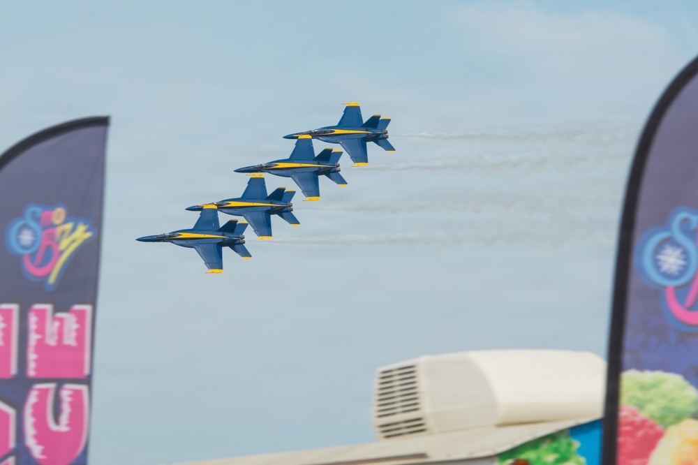 Four U.S. Navy Blue Angels jets fly in a tight diamond formation against a pale blue sky, leaving white smoke trails behind them. The aircraft are painted in their distinctive blue and gold livery. The scene is captured from ground level at what appears to be an airshow, with vendor banners visible in the foreground.