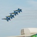Four U.S. Navy Blue Angels jets fly in a tight diamond formation against a pale blue sky, leaving white smoke trails behind them. The aircraft are painted in their distinctive blue and gold livery. The scene is captured from ground level at what appears to be an airshow, with vendor banners visible in the foreground.