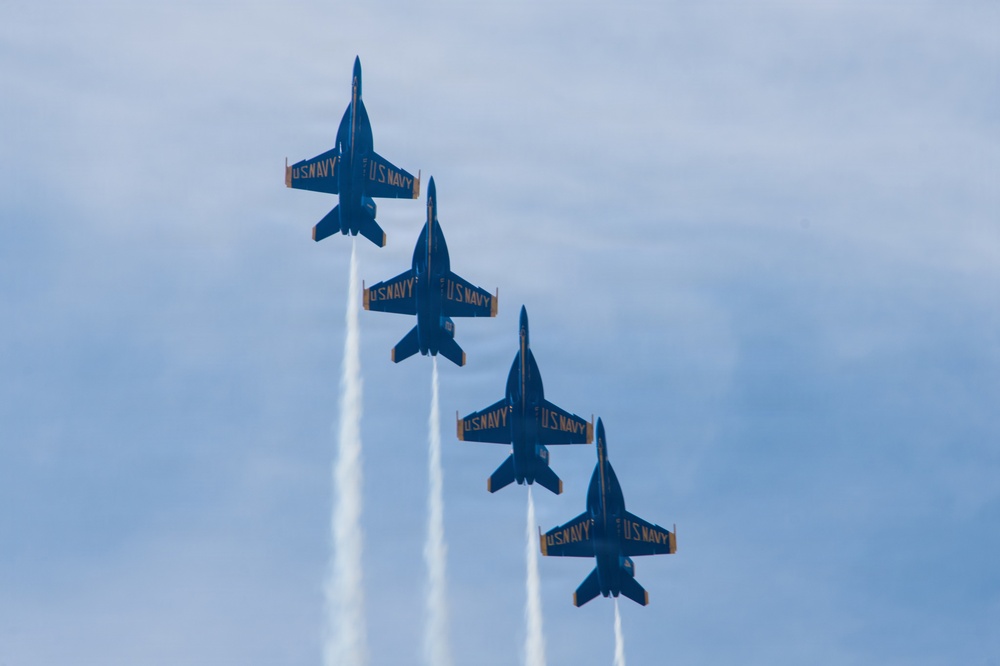 Four U.S. Navy Blue Angels jet aircraft are flying in a diagonal echelon formation, viewed from below against a partly cloudy blue sky. Each aircraft is producing a white smoke trail. The jets are painted in the distinctive blue and gold color scheme with 'U.S. NAVY' markings visible on the wings.