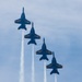 Four U.S. Navy Blue Angels jet aircraft are flying in a diagonal echelon formation, viewed from below against a partly cloudy blue sky. Each aircraft is producing a white smoke trail. The jets are painted in the distinctive blue and gold color scheme with 'U.S. NAVY' markings visible on the wings.