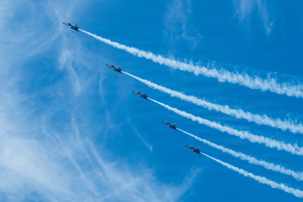Five military jet aircraft fly in a tight diagonal echelon formation against a vivid blue sky, each producing a distinct white smoke trail. The aircraft appear to be performing an aerobatic demonstration, consistent with a precision flight team such as the U.S. Navy Blue Angels. The parallel smoke trails create a striking visual pattern extending from upper-left to lower-right across the frame.