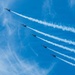 Five military jet aircraft fly in a tight diagonal echelon formation against a vivid blue sky, each producing a distinct white smoke trail. The aircraft appear to be performing an aerobatic demonstration, consistent with a precision flight team such as the U.S. Navy Blue Angels. The parallel smoke trails create a striking visual pattern extending from upper-left to lower-right across the frame.