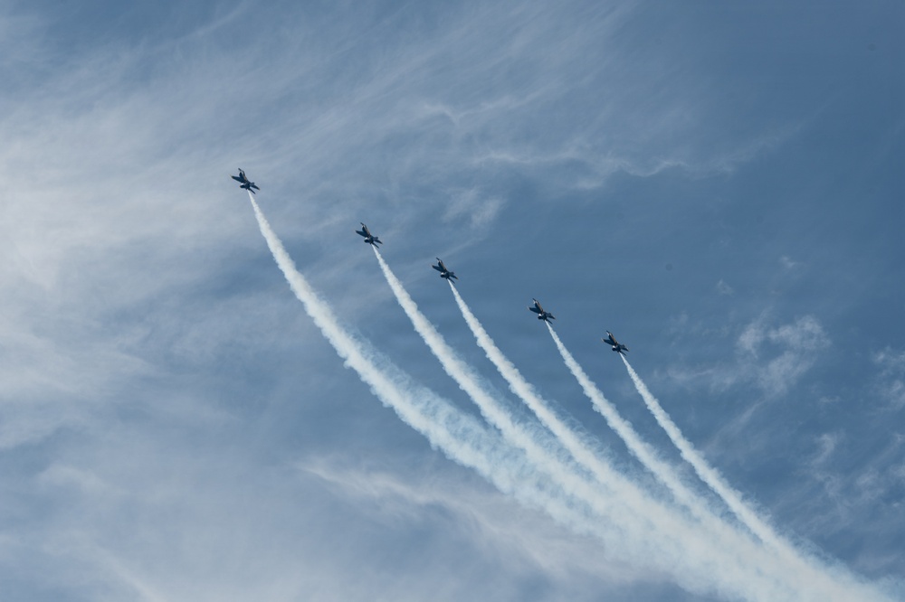 Five military jet aircraft are flying in a diagonal echelon formation against a partly cloudy blue sky. Each aircraft is producing a white smoke or contrail trail behind it, creating a visually striking parallel pattern. The formation appears to be part of an aerobatic or ceremonial air show display.