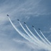 Five military jet aircraft are flying in a diagonal echelon formation against a partly cloudy blue sky. Each aircraft is producing a white smoke or contrail trail behind it, creating a visually striking parallel pattern. The formation appears to be part of an aerobatic or ceremonial air show display.