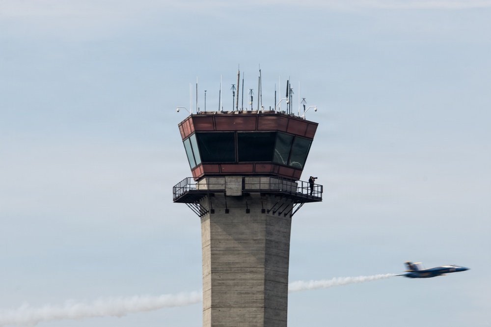 The image shows an airport air traffic control tower constructed of concrete with a glass-enclosed cab at the top, featuring a wraparound observation deck. A person is visible on the exterior walkway of the tower, appearing to photograph or observe. In the background to the right, a blue and yellow jet aircraft — consistent with a U.S. Navy Blue Angels demonstration team aircraft — is flying past leaving a white smoke trail, suggesting an airshow is in progress.