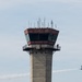 The image shows an airport air traffic control tower constructed of concrete with a glass-enclosed cab at the top, featuring a wraparound observation deck. A person is visible on the exterior walkway of the tower, appearing to photograph or observe. In the background to the right, a blue and yellow jet aircraft — consistent with a U.S. Navy Blue Angels demonstration team aircraft — is flying past leaving a white smoke trail, suggesting an airshow is in progress.