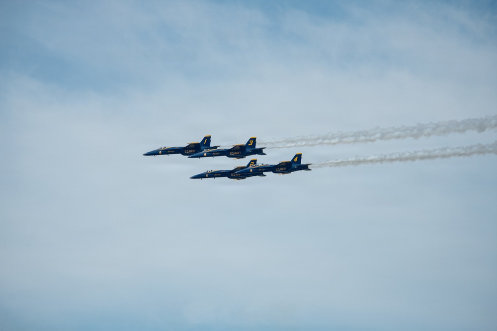 Four U.S. Navy Blue Angels aircraft are flying in a tight diamond formation against a partly cloudy blue sky. The jets, numbered 1 through 4, are producing white smoke trails as they perform at what appears to be an airshow. The aircraft are painted in the iconic blue and gold Blue Angels livery.