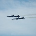 Four U.S. Navy Blue Angels aircraft are flying in a tight diamond formation against a partly cloudy blue sky. The jets, numbered 1 through 4, are producing white smoke trails as they perform at what appears to be an airshow. The aircraft are painted in the iconic blue and gold Blue Angels livery.