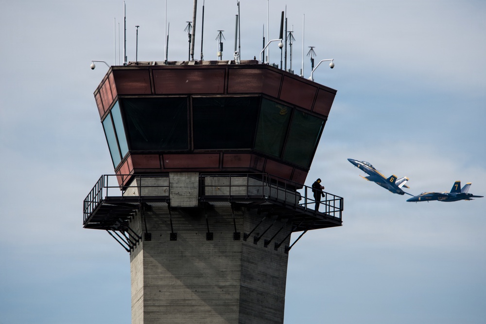 The image shows an air traffic control tower with a person standing on its exterior observation walkway, watching two military jet aircraft flying in the background. The jets are painted in the distinctive blue and gold color scheme of the U.S. Navy Blue Angels flight demonstration squadron, with visible numbers '6' and '7' on their fuselages. The scene appears to be captured during an airshow or flight demonstration event.