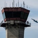 The image shows an air traffic control tower with a person standing on its exterior observation walkway, watching two military jet aircraft flying in the background. The jets are painted in the distinctive blue and gold color scheme of the U.S. Navy Blue Angels flight demonstration squadron, with visible numbers '6' and '7' on their fuselages. The scene appears to be captured during an airshow or flight demonstration event.