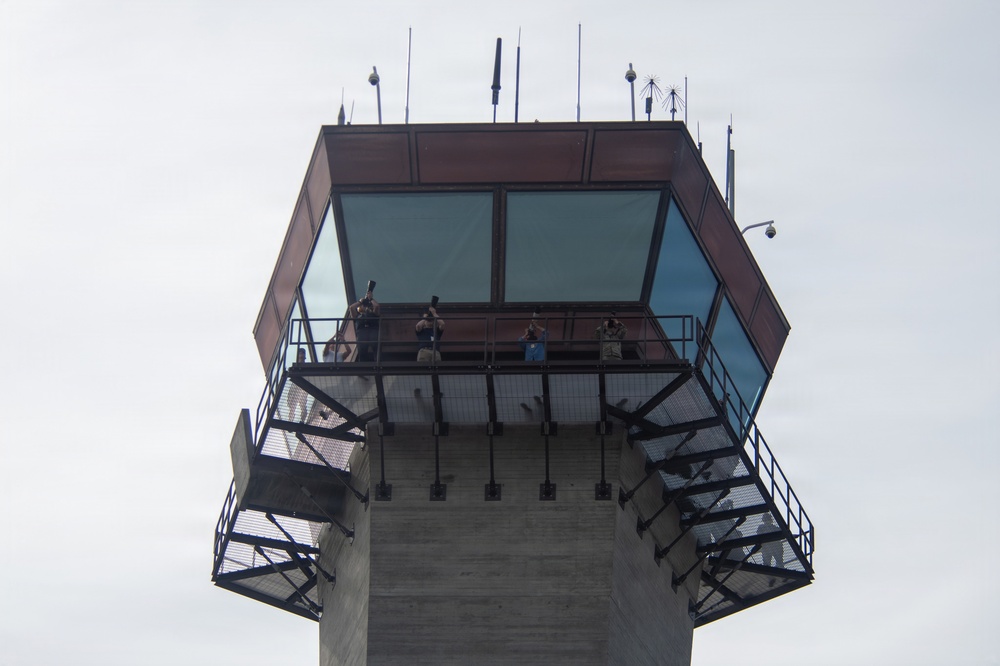 The image shows the upper portion of what appears to be an air traffic control tower, photographed from a low angle looking upward. The tower features a glass-enclosed cab at the top with an exterior observation walkway, where several people are visible. Various antennas, communication equipment, and a security camera are mounted on the roof.