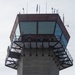 The image shows the upper portion of what appears to be an air traffic control tower, photographed from a low angle looking upward. The tower features a glass-enclosed cab at the top with an exterior observation walkway, where several people are visible. Various antennas, communication equipment, and a security camera are mounted on the roof.