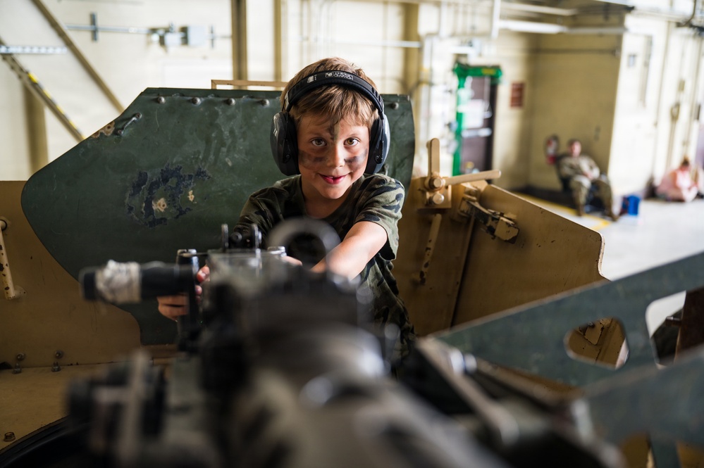 A young boy wearing hearing protection headphones and camouflage face paint is seated behind what appears to be a military vehicle-mounted weapon or equipment, smiling at the camera. He is dressed in a camouflage t-shirt, suggesting participation in a military-themed event or open day. The setting appears to be an indoor military hangar or facility with other people visible in the background.