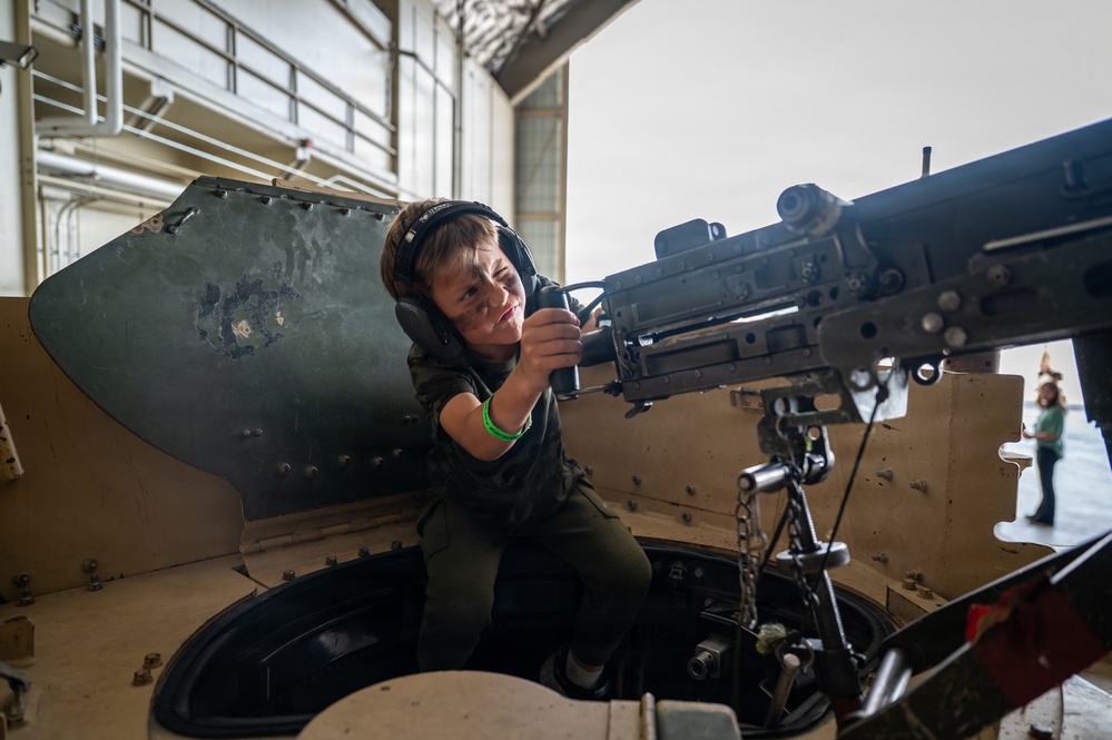 A young child wearing ear protection headphones and a green wristband is seated in the turret hatch of a military armored vehicle, gripping a mounted machine gun. The setting appears to be an indoor military hangar or exhibition facility, suggesting this is a public open day or air show event. The child is smiling, indicating this is a supervised interactive display rather than an operational context.